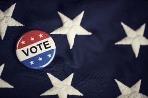A red, white, and blue button that says "VOTE" lies atop the starry blue field of an American flag.
