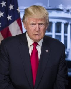 A serious looking Donald Trump scowls in front of a display of the American Flag and the White House.