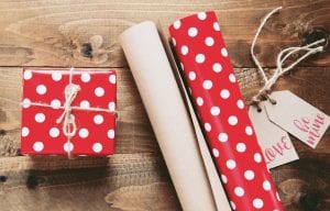 Box wrapped in red paper with white dots sitting next to wrapping paper and gift tags saying &ldquo;Love&rdquo; and &ldquo;Be mine.&rdquo; Image by Miroslava, via Unsplash.com.