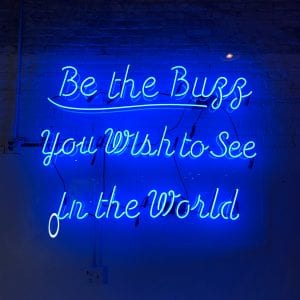 Neon blue &ldquo;Be the buzz you wish to see in the world&rdquo; sign; image by Tanya Santos, via Unsplash.com.