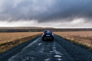 Car driving down dirt road through fields of brown grass, storm clouds in the sky; image by K&eacute;vin Langlais, via unsplash.com.