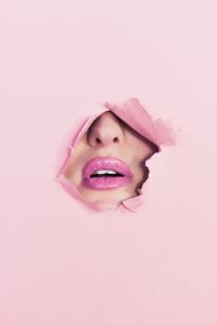 Woman&rsquo;s nose and mouth, visible through a hole torn in pink paper background; image by Ian Dooley, via unsplash.com.