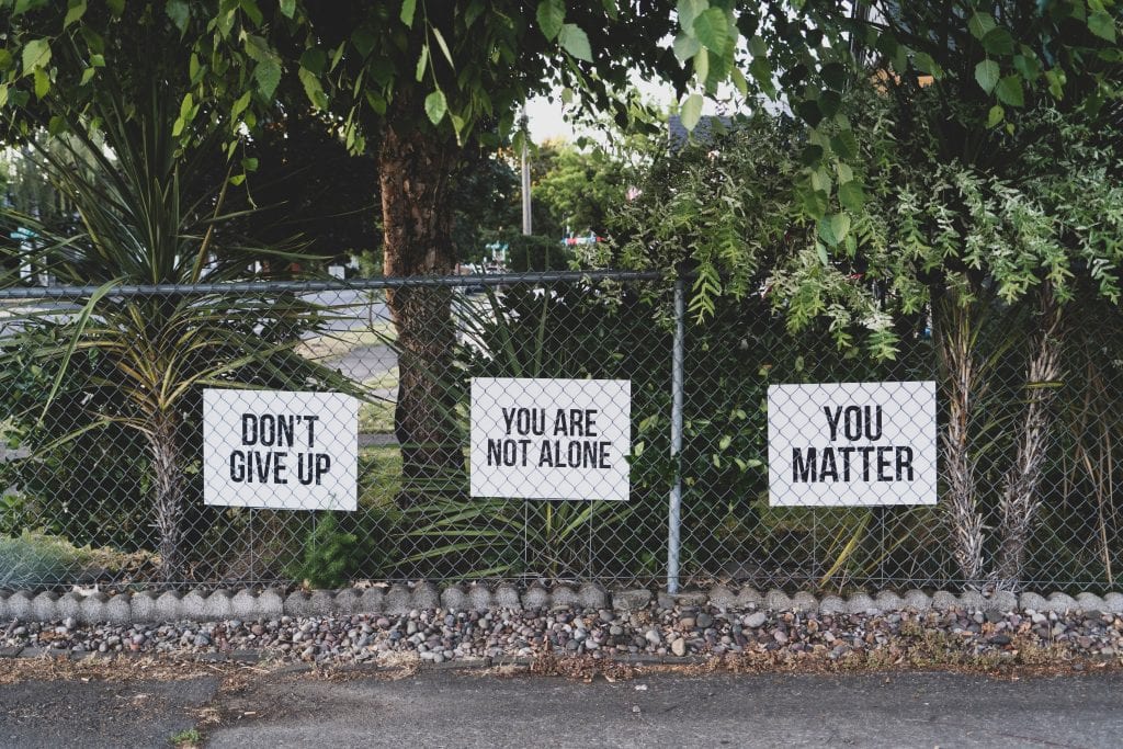 Three signs on a chain link fence, reading (in order) &ldquo;Don&rsquo;t Give Up,&rdquo; &ldquo;You Are Not Alone,&rdquo; and &ldquo;You Matter.&rdquo; Image by Dan Meyers, via Unsplash.com.