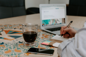 Person writing, with cellphone and coffee nearby; image by Kaleidico, via Unsplash.com.