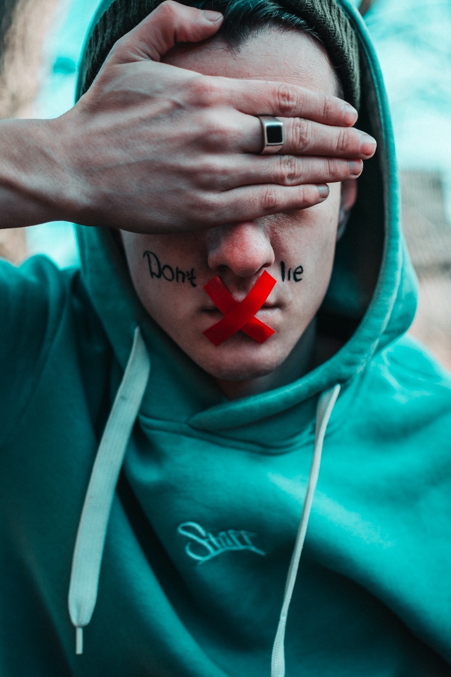Man with his hand covering his eyes, red tape in an X on this mouth, and the words "Don't Lie" written on either side of his nose; image by Taras Chernus, via Unsplash.com.
