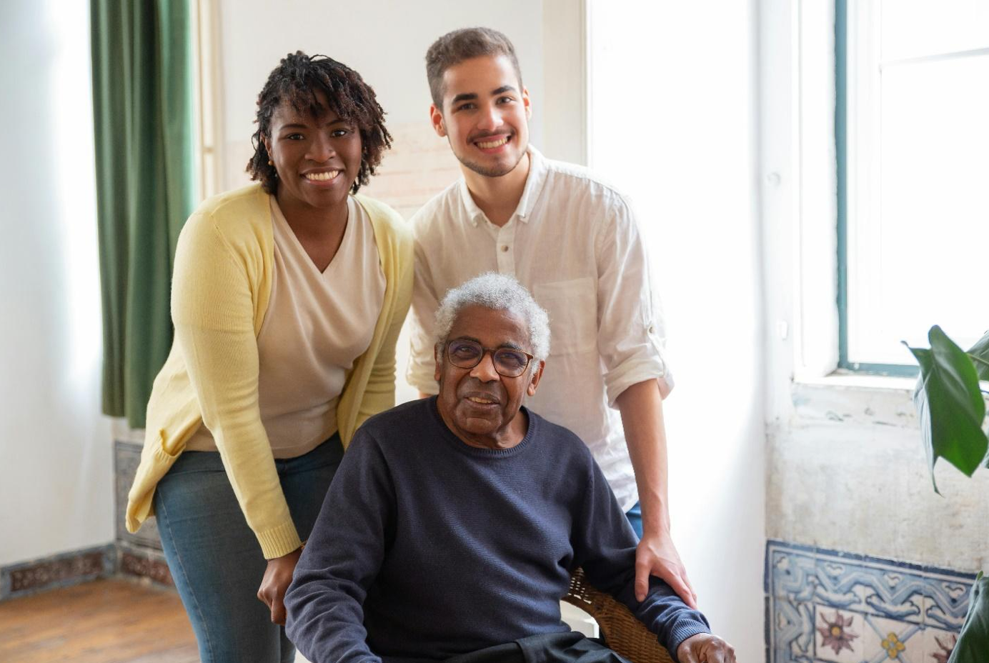 Man in wheelchair, two people standing behind him. Everyone is smiling. Image by Kampus Production, via Pexels.com.